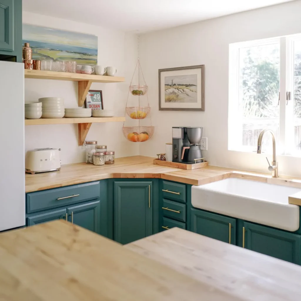 A beautiful Kitchen with Butcher block countertop with a sink fitted in it, jars, some kitchen appliances on countertop. Shelves with cups, glasses and plates. A hanging fruit basket along the wall.