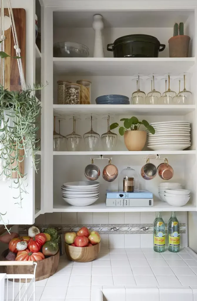 A beautiful kitchen with white tile countertop with some fruit and vegetables in baskets. There are various type of utensils in shelves 