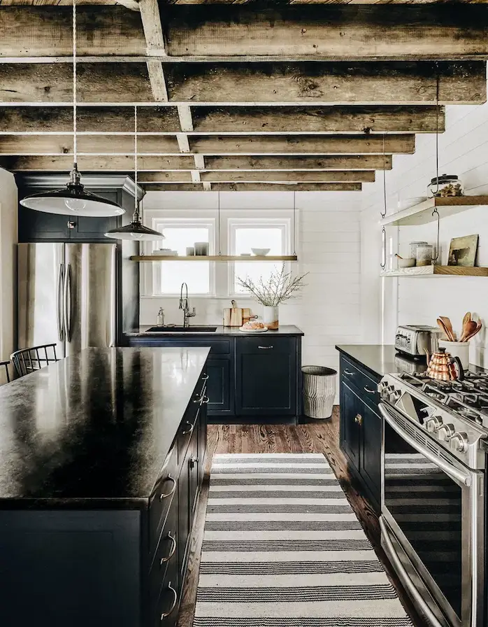 A modern kitchen with cabinets in a semi-gloss blue-black color and The black worktops with the white shiplap walls, rustic wood floors, and exposed ceiling beams